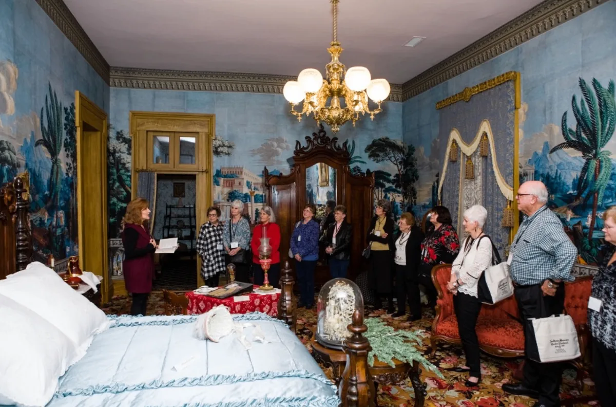 Tamera Alexander leading guests through a blue historic bedroom during the Southern Mansion Reader Weekend