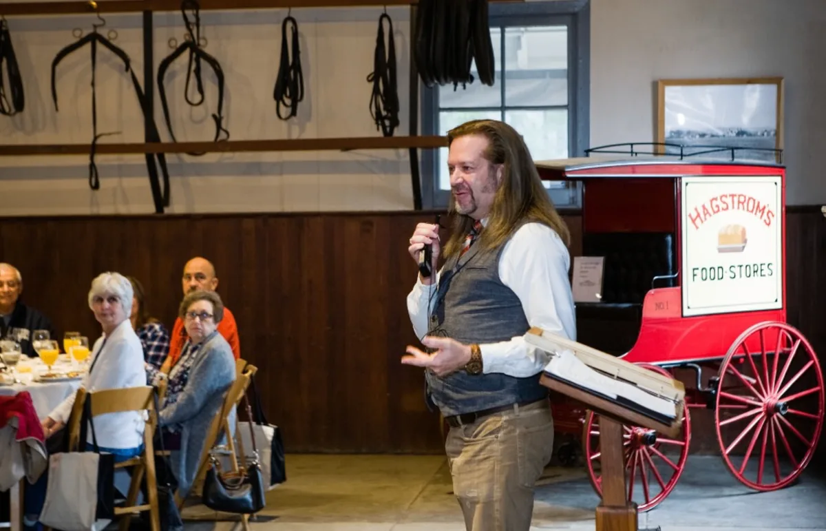 A speaker addressing Southern Mansion Reader Weekend attendees beside a red historic wagon